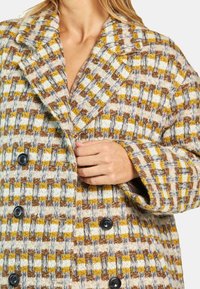 Dwurzędowy płaszcz z tkaniny w plecioną kratę w odcieniach brązu, żółci i bieli. Posiada oversize'owe klapy oraz duże czarne guziki. Teksturowana tkanina.