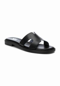 Sandalia de piel negra con dos correas anchas que presentan acentos en forma de corte, suela plana y un diseño minimalista.