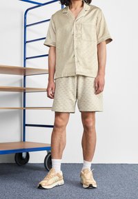 Ljusbeige pyjamasset som består av en kortärmad skjorta med knappar och shorts med ett subtilt bladmönster. Bärs tillsammans med tanfärgade sneakers.