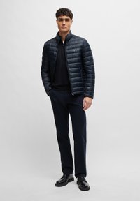 Marineblauwe pufferjack met horizontale stiksels, hoge kraag en ritssluiting; gecombineerd met een zwarte trui, marineblauwe broek en zwarte schoenen.