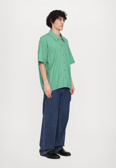 Green checkered short-sleeve shirt with a relaxed fit, paired with loose blue denim jeans and black shoes.