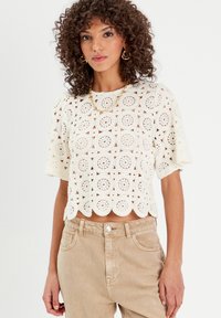 Top blanc court avec des motifs de dentelle circulaires et un ourlet festonné. Des manches courtes évasées complètent le design complexe, fabriqué à partir d'un tissu léger.