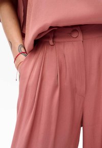 Różowe spodnie z zakładkami, z zapięciem na guzik w pasie i kieszeniami bocznymi. Tkanina wydaje się gładka z lekkim połyskiem. Brak widocznych wzorów.