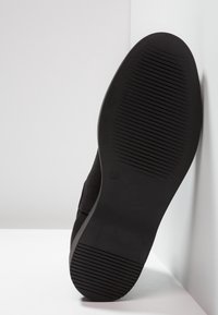 Semelle de chaussure noire avec des rainures horizontales au niveau du talon et de l'avant-pied, inclinée contre un mur et une surface blancs.