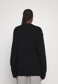 Person mit geflochtenem Haar, der einen lässigen schwarzen Strickpullover und hellgraue Hosen trägt und mit dem Rücken zu einem schlichten hellen Hintergrund steht.
