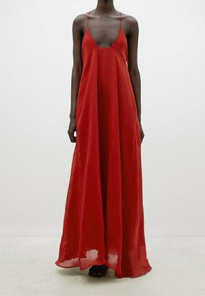 Femme portant une longue robe rouge fluide sans manches avec de fines bretelles, debout devant un fond clair uni.