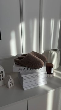 Pruunid karvased sussid, millel on paks ja tekstureeritud pind, paiknevad virnastatud raamatute peal. Lähedal on valge sooniline vaas ja küünal heledal pinnal.