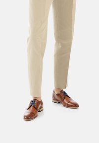 Brązowe skórzane buty eleganckie o gładkiej fakturze, z niebieskimi sznurówkami i okrągłym noskiem, połączone z jasnobeżowymi spodniami.