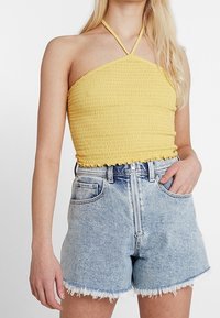 Žena na sobě má žlutý halter top s nabíraným efektem, který je spojený s vysokým pasem světle modrými džíny šortkami s předním zipem a roztrhanými lemy.