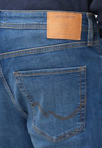 Pantalones vaqueros de mezclilla en azul medio con costuras naranjas, un bolsillo trasero y un parche de marca de cuero marrón con texto en relieve.