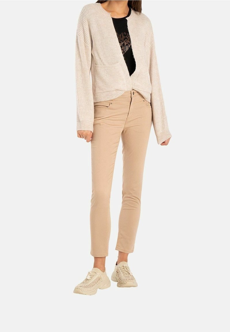 Cardigan côtelé beige clair avec des manches longues, une poche avant, associé à un pantalon beige clair ajusté et des baskets crème.