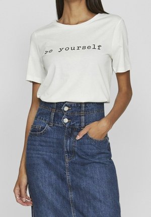 Femme portant un t-shirt blanc à manches courtes avec le texte « sois toi-même » et une jupe en denim bleu taille haute avec fermeture à double bouton, main dans la poche.