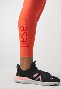 Oransje tights med glinsende "Casall" logo detalj på siden og svarte treningssko med rosa detaljer og slip-on design.