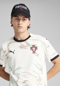 Biała koszulka sportowa z czerwonymi i zielonymi wzorami, z herbem piłkarskim Portugalii i logo PUMA. Czarna czapka z napisem "PORTUGAL".