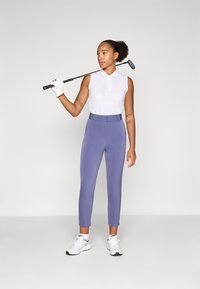Polo sans manches blanc avec col, associé à un pantalon de golf lavande ajusté et des chaussures de sport blanches, tenant un club de golf.