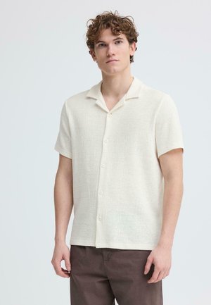 Jonge man met krullend haar, gekleed in een kort mouwens overhemd in roomkleur en een bruine broek, staand tegen een effen lichte achtergrond.