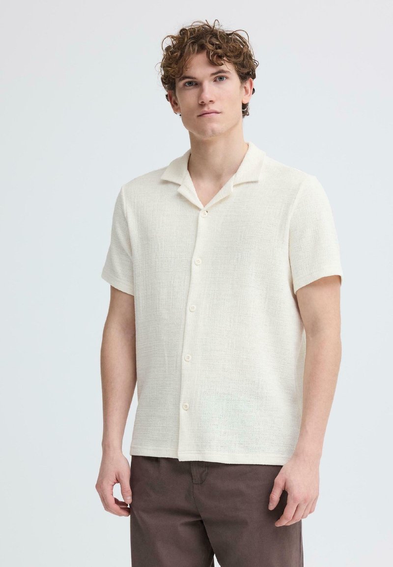 Mladý muž s kučeravými vlasmi, oblečený v krémovej košeli s krátkym rukávom a hnedých nohaviciach, stojaci pred jednoduchým svetlým pozadím.