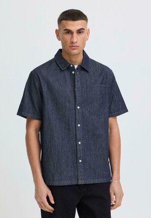 Ung mand med kort hår iført mørkeblå kortærmet denimskjorte med knapper og brystlomme, stående mod en ensartet lys baggrund.