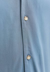 Jasnoniebieska tkanina z dwoma przyszytymi beżowymi guzikami ułożonymi pionowo na listwie koszuli.