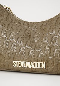 Brun struktureret håndtaske med prægede logo mønster og gyldne detaljer. Har en lynlåslukning og viser tydeligt "STEVE MADDEN" i guld.