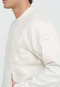 Felpa beige chiara con scollo a girocollo, maniche raglan, tessuto texturizzato e un discreto logo applicato sulla manica. Presenta una grande tasca frontale.