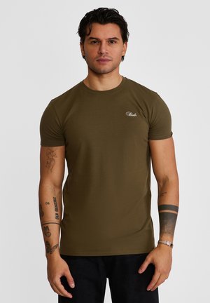 Mężczyzna o krótkich ciemnych włosach, ubrany w oliwkowo-zielony dopasowany T-shirt i czarne spodnie, z wieloma tatuażami na obu ramionach, stoi na tle jednolitego tła.