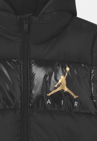 Sort oppustet jakke med skinnende tekstur, prydet med et gyldent Jordan-logo og hvid "AIR" tekst på forsiden. Lynlås-lukning og hætte-design.