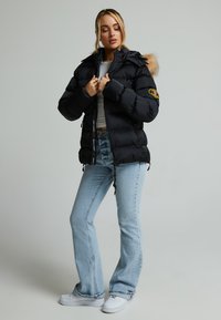 Svart puffer-jakke med avtakbar pelshette, sidelommer og logo-patch på ermet, kombinert med lyseblå jeans og hvite joggesko.