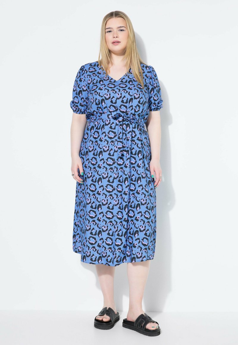 Vestido de estampado de leopardo azul con escote en V, mangas cortas enrolladas y cintura ceñida con cinturón. Hecho de una tela liviana, cae hasta la mitad de la pantorrilla. Sandalias negras.