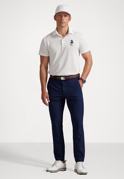 Bărbat îmbrăcat cu o șapcă albă de golf, un polo alb cu logo-ul unui urs, pantaloni bleumarin, o curea maro, un ceas la încheietură și încălțări albe de golf, stând în fața unui fundal simplu.