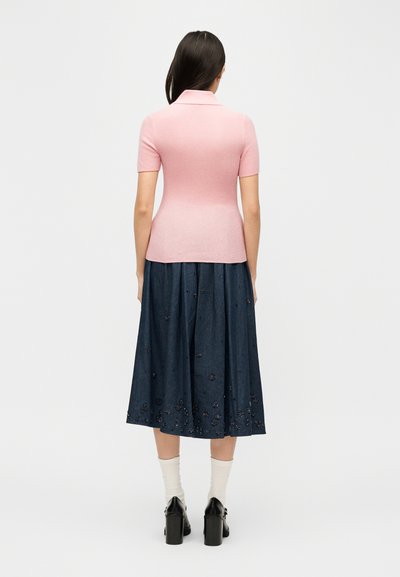 Γυναίκα που στέκεται με την πλάτη προς τα έξω, φοράει στενό ροζ κοντομάνικο τοπ, μεσομήκη μπλε φούστα με κέντημα, λευκές κάλτσες και μαύρα παπούτσια με τακούνι.
