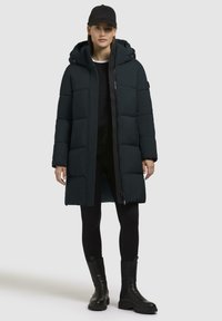 Palton puffer negru, căptușit, cu glugă, fermoar frontal și buzunare laterale, având o textură netedă și un design până la genunchi. Purtați cu colanți negri și cizme.