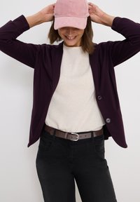 Mörklila blazer med två knappar över en ljusbeige tröja, kombinerat med svarta byxor och en rosa kordflöjtskeps. Enkla designfunktioner.