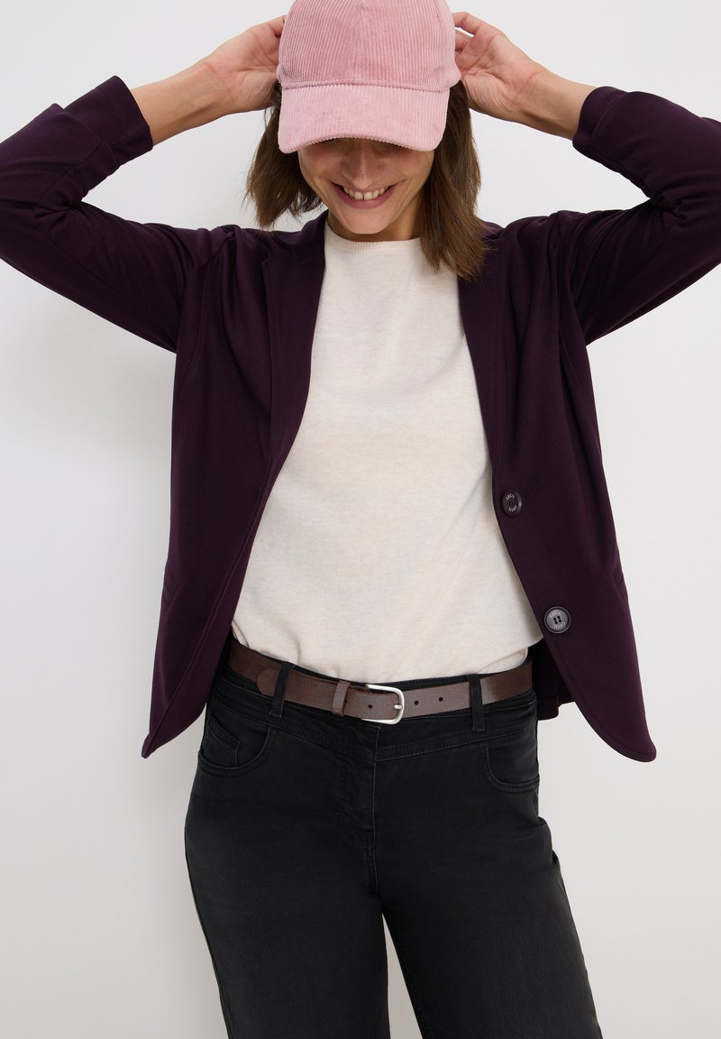 Mörklila blazer med två knappar över en ljusbeige tröja, kombinerat med svarta byxor och en rosa kordflöjtskeps. Enkla designfunktioner.