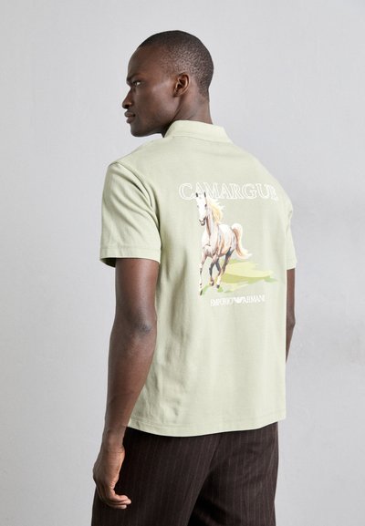 Svijetlozelena pamučna polo majica s ilustracijom konja na leđima, s natpisom "CAMARGUE" i logom "Emporio Armani" ispod.
