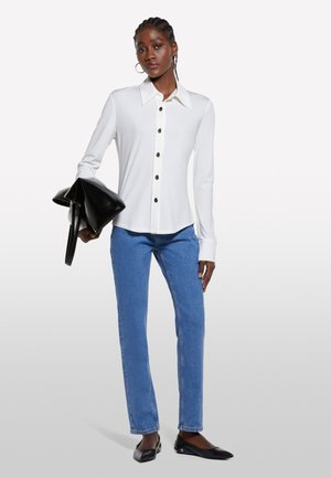 Camicia bianca a maniche lunghe con colletto a punta e bottoni marroni, abbinata a jeans blu a gamba dritta e ballerine nere, mentre tiene una borsa nera.