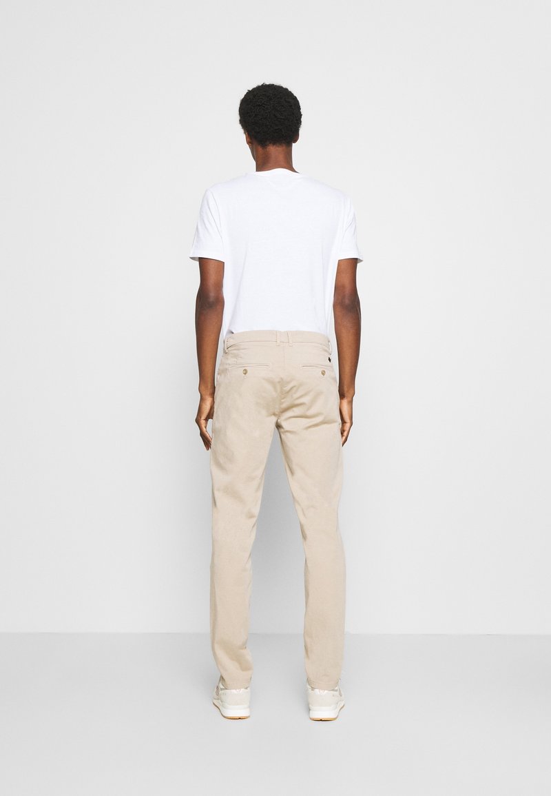 Homme vu de dos, portant un t-shirt blanc à manches courtes, un pantalon beige et des baskets blanches, debout devant un fond uni gris clair.