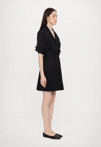 Rochie neagră cu curea, cu mâneci scurte, decolteu în V și croi drept, purtată cu balerini negri. Textură netedă și design minimalist.