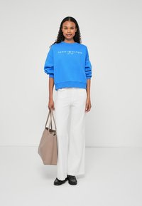 Modrá mikina s bielym logom, biele široké nohavice, čierne topánky a béžová taška. Model stojí proti neutrálnemu pozadiu.