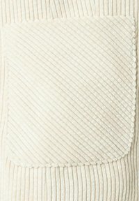 Crème pluche stof met diagonale ribbelstructuur; beschikt over een vierkante zak met golvende randen. Het ontwerp benadrukt zachtheid en warmte.