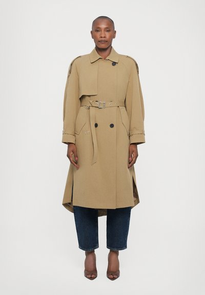 Beež trench coat, millel on vööga talje, topelt-nupud ja suured taskud, kihtide kaupa siniste lühikeste teksade ja pruunide kontsade peal.