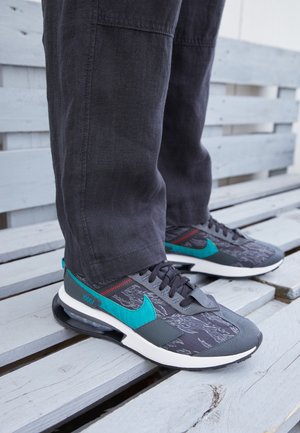 Sneakers neri, grigi e teal con sovrapposizioni testurizzate, dotati di una suola imbottita e dettagli rossi. Indossati con pantaloni scuri.