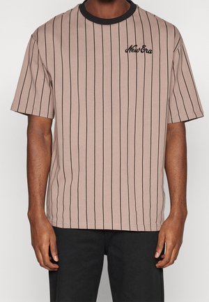 Mann trägt ein beiges T-Shirt mit kurzen Ärmeln, schwarzen vertikalen Streifen, schwarzem Kragen und dem Schriftzug "New Era" auf der linken Brust gestickt.