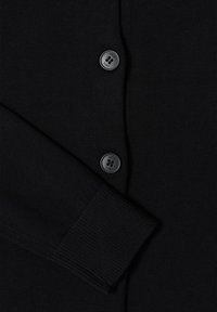 Manche et devant d'un cardigan en maille noire avec deux boutons noirs gravés du mot « CECIL » sur un tissu texturé.