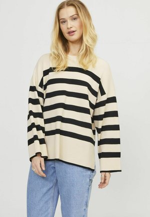 Maglioncino leggero a righe beige e nero, con maniche lunghe, texture a coste e taglio ampio. Abbinato a jeans di denim azzurri.