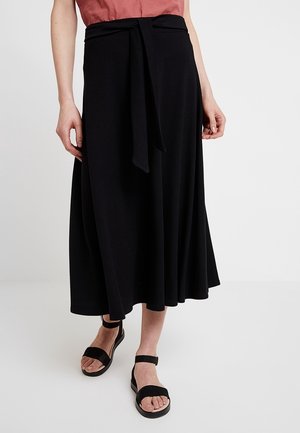 Zwarte midilange rok van zachte stof. Voorzien van een taille met riem en een A-lijn silhouet. Draagt zwarte sandalen.