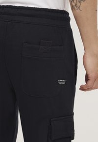 Zwarte joggingsbroek met een zijcargo pocket, een grote achterzak, een elastische tailleband en een merklabel op de achterzak.