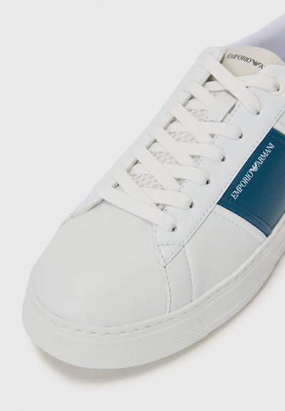 Vit lädersneaker med blå detaljer, rund tå och gummisula. Har platta snören och "Emporio Armani"-logotyp på sidan.