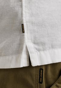Camisa de lino blanca con una abertura lateral, que presenta una etiqueta marrón. La textura y las costuras visibles contrastan con los pantalones verde oliva que se encuentran debajo.