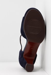 Une seule chaussure en daim bleu marine reposant sur le côté montrant la semelle et le talon marron avec des zones de grip texturées et le nom de la marque estampillé.